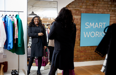 A young Black woman tries on some office clothes in front of a full-length mirror, smiling. On the wall next to the mirror is a sign saying Smart Works.