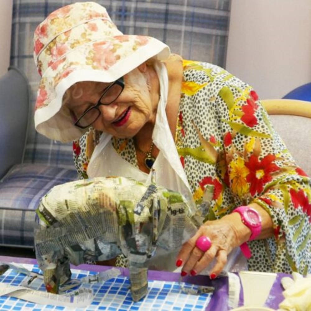 An elderly lady wearing bright floral clothes tends to a papier mache elephant model indoors