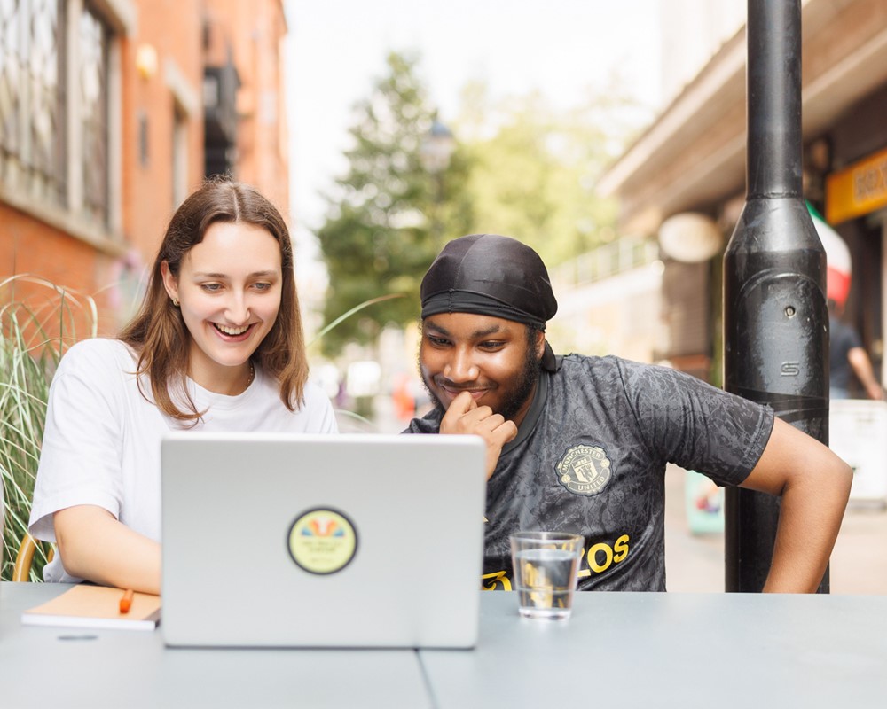 Two young people of different ethnicities sit together at an outdoor table on a walkway smiling and looking at an open laptop together