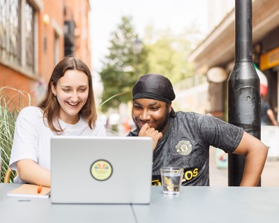 Two young people of different ethnicities sit together at an outdoor table on a walkway smiling and looking at an open laptop together