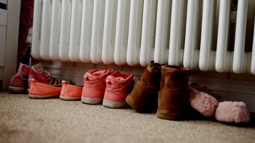 A row of shoes of various sizes and types are huddled in a hallway under a radiator.