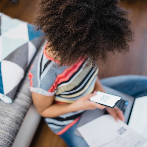 A young black woman sits on a laminated floor against a sofa cross legged, holding a phone in one hand and a piece of paper in the other, looking at the laptop balanced on her lap.