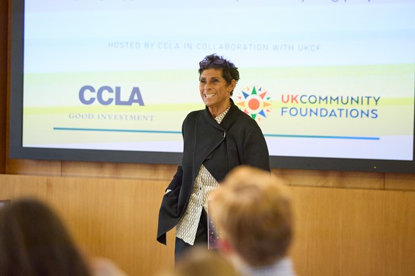 Fatima Whitbread MBE stands smiling in front of an audience at UKCF conference, hosted by CCLA, with a big screen behind her.