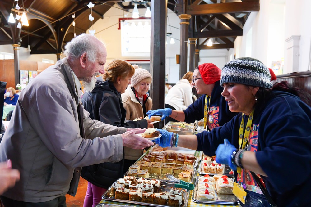 Volunteers in a church hall pass out sandwiches and cake across a busy counter to a mix of elderly and younger people all eager to have their lunch.