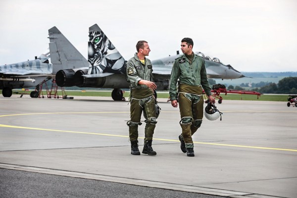 Photo of Ajvir Singh Sandhu walking along a runway with a co-pilot under a cloudy sky. Ajvir is wearing a khaki air cadet uniform, holding a helmet and surrounded by fighter jets.