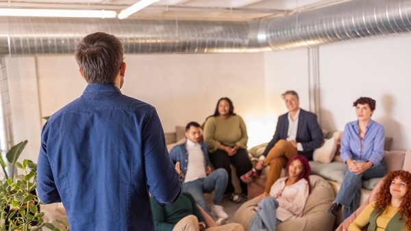 A meeting taking place in a light room where 6 people in casual clothes rest on beanbags while listening to a person speaking in front of them.