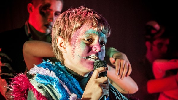 Photo of a young person on stage at night with young dancers around them. The young person in focus has face paint on in a colourful costume and is holding a microphone.