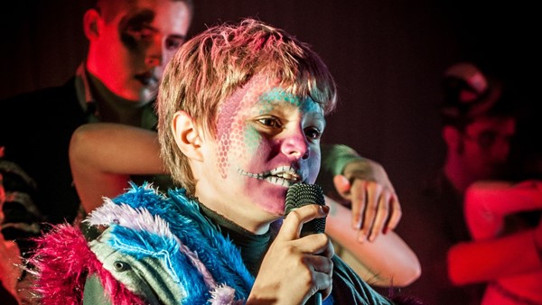 Photo of a young person on stage at night with young dancers around them. The young person in focus has face paint on in a colourful costume and is holding a microphone.