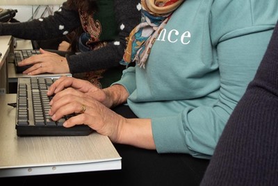 Two women, one of whom is wearing a colourful hijab and looking down smiling, are sitting in front of computers and typing.