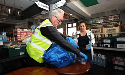 A male volunteer and a female volunteer, both wearing hi-vis jackets, pack food containers into bags inside a pub converted into a food hub