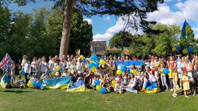 Ukrainian families and individuals sit outside in a field on a sunny day, waving Ukrainian flags and yellow and blue balloons.