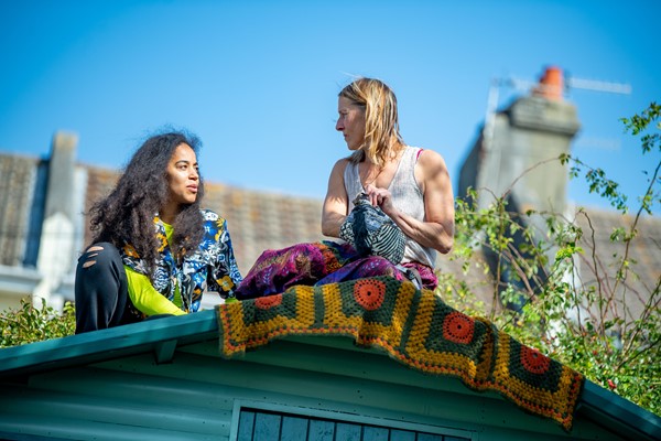 Two women of different cultural heritages sit upon a shed roof talking to each other in the sunshine.
