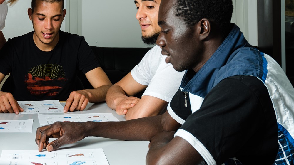A group of men of different nationalities are sat around a large table taking part in a learning activity with worksheets