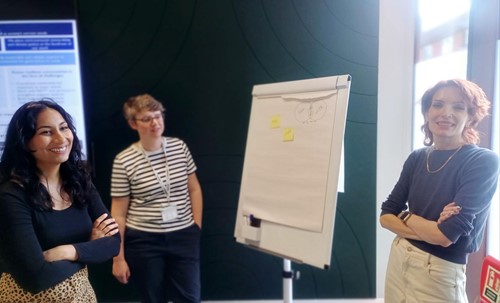 Three women of different ethnicities stand together around a whiteboard in an office, smiling, having added written post-it notes to the board.