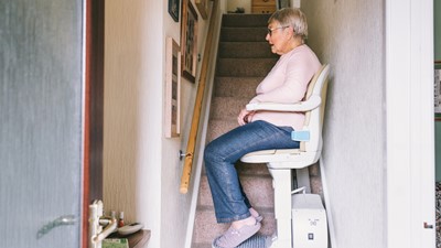 Older woman sits in a stairlift in her home mid-stairs, in jeans, a jumper and slippers. 