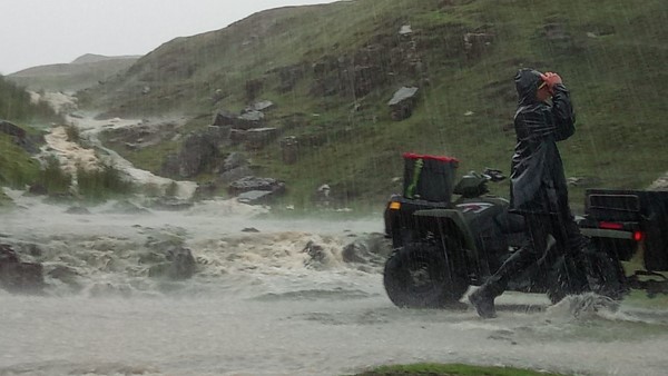 A person wearing a raincoat hols their hood over their face firmly as they walk past a mini tractor in a rugged landscape while it rains heavily on soaked land.