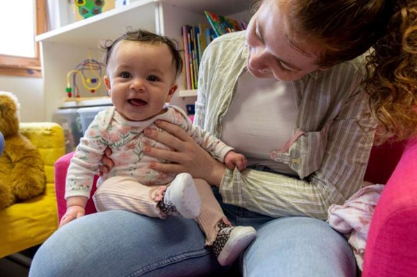 A young mum bounces a very happy baby on her knee while seated on the floor surrounded by toys and books
