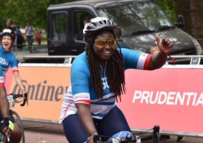 A Black woman in lycra cycles towards a crowd with her thumb up to them and a big grin during a road race with other contestants in the background