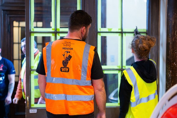 A mentor in a high-vis jacket stands talking with a young volunteer in a high-vis in front of a warehouse window 