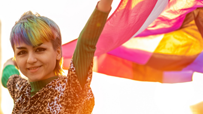 Photo of a young non-binary person holding a multicoloured flag that symbolises trans equality