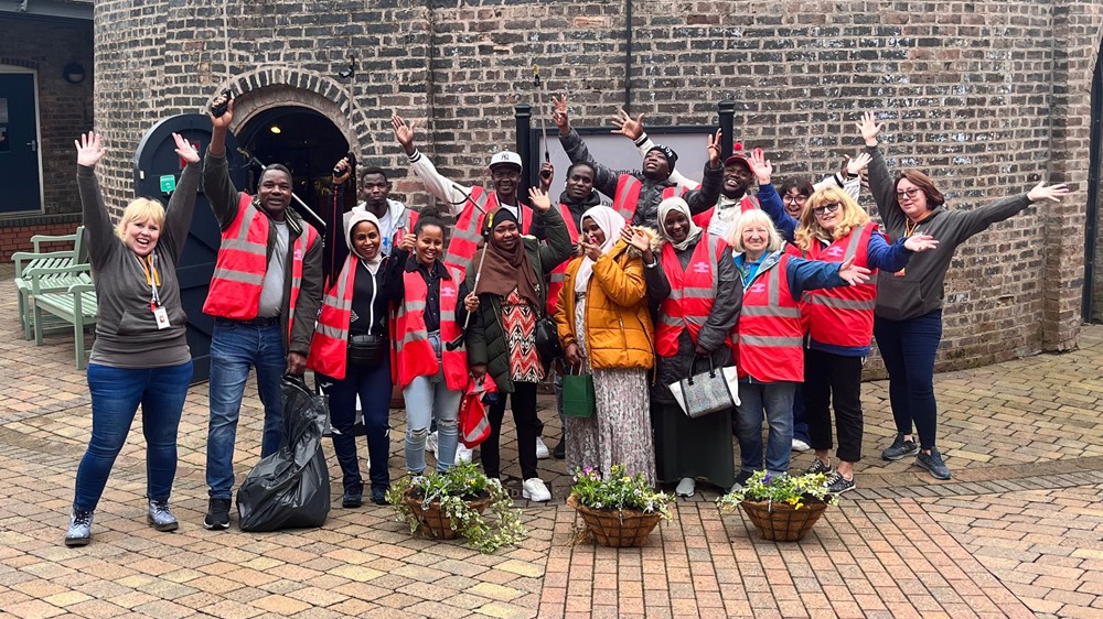 A group of 16 people of mixed ethnicities stand in a row outside an old building with high vis vests on, holding litter pickers and bin bags, smiling and waving. 