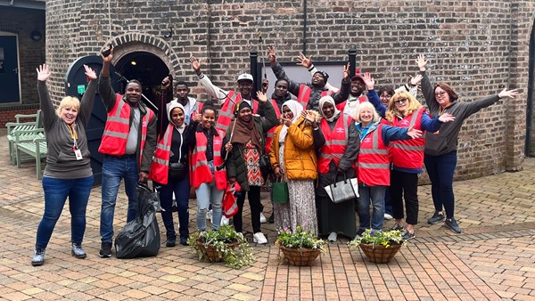 A group of 16 people of mixed ethnicities stand in a row outside an old building with high vis vests on, holding litter pickers and bin bags, smiling and waving. 