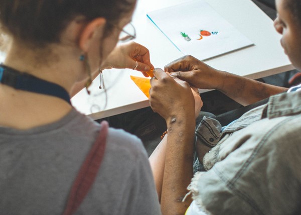 Two women of different ethnicities work on the same piece of origami together in deep concentration at a table in a workshop.